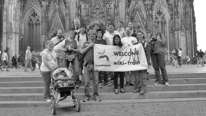 Un groupe de Wikimédiens et Wikimédiennes posant devant la cathédrale de Cologne avec une bannière au logo Wikimania disant "Welcome wiki-train"