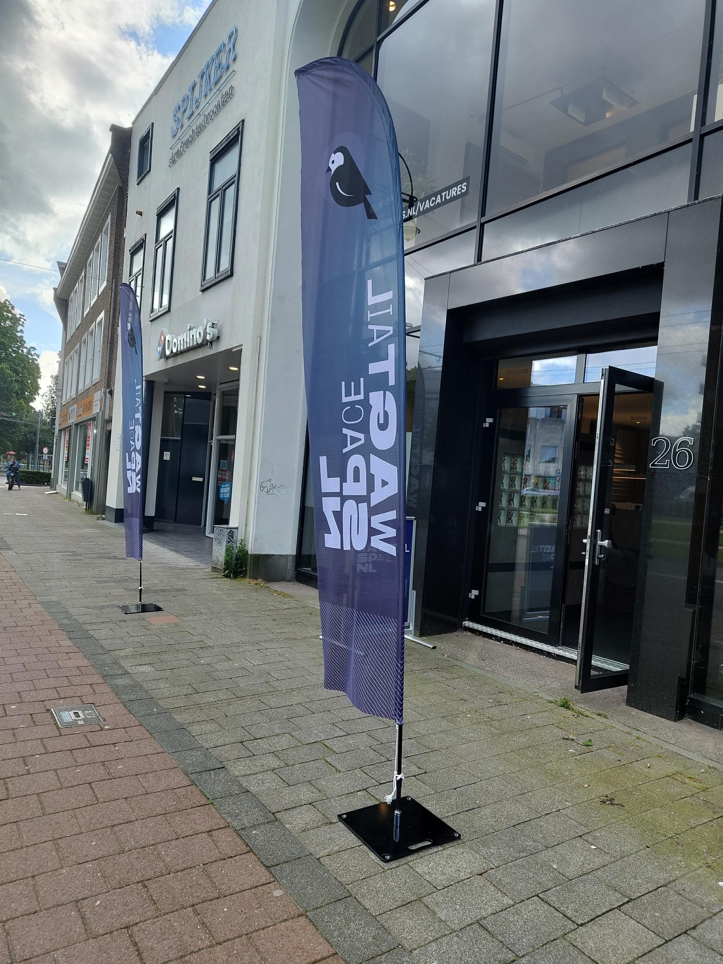 La porte d'un immeuble de bureaux vitré, encadrée par deux drapeaux de type flamme bleus avec le logo de Wagtail (une bergeronnette noire et blanche) surmontant la mention "Wagtail Space NL" en vertical et en capitales.